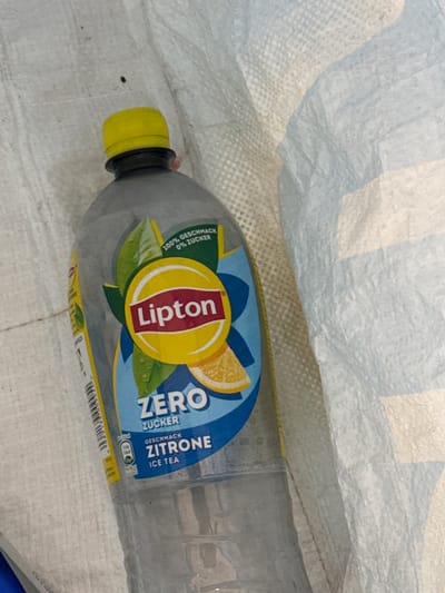A clear plastic bottle of Lipton Zero Citrone Ice Tea is lying on a white, textured surface, likely a woven bag or tarp. The bottle has a yellow cap and features a label with the Lipton logo, a stylized sun with the brand name in red, and graphics of green leaves and a slice of lemon. Text on the label indicates "100% Geschmack 0% Zucker" (100% flavor 0% sugar) and "Geschmack Zitrone Ice Tea" (Lemon flavor Ice Tea). The scene appears to be outdoors or in a storage area, and no people are present. The location in Oosterhout, Netherlands, is not ascertainable from the image.