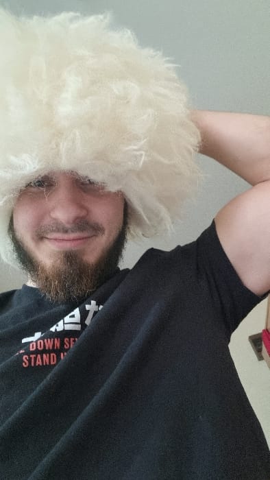 The image displays a waist-up portrait of a single male individual. The subject has light skin, dark eyes, and a prominent dark brown beard and mustache, and is smiling directly at the camera. He is wearing a large, shaggy, off-white or cream-colored hat that covers his forehead and the upper part of his head. He is dressed in a black short-sleeved t-shirt with partial white and orange text, from which the words "DOWN SE" and "STAND" are discernible. His right arm is bent at the elbow, with the hand positioned behind his head or hat, partially obscured. The background is a plain, light-colored interior wall, possibly white or light grey. A small fragment of a red object or fabric is visible in the lower right corner of the frame. The specific location within the city of Belozernyi, Russia, cannot be determined from the visual information provided in the image.