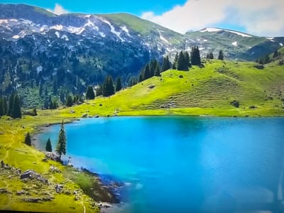 The image shows a mountain landscape with a blue lake in the foreground. The mountains are green with patches of snow. There are scattered pine trees on the hillside. The sky is blue with some clouds. The location is not in Basingstoke and Deane, United Kingdom.