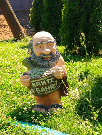 A garden gnome statue stands in a grassy area with small white flowers. The gnome is bald with a bushy beard and is holding a round object with writing on it. The writing is in Cyrillic script and appears to say "КАЙТЕ В БАНЮ!" which translates to "Go to the bathhouse!". The gnome is wearing a brown garment over its shoulders and its feet are visible at the bottom of the statue. Behind the gnome, there are green evergreen trees. The ground is covered in green grass and small white and yellow flowers. In the background, there is a wooden post and a pile of brown dirt. The scene is brightly lit by the sun. The location is the city of Kok Jar, Kyrgyzstan.