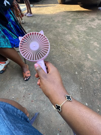 In Amurie, Nigeria, a person's hand holds a pink, cat-eared portable fan. The fan's circular head features radiating lines and a simple cat face design. The person's arm is adorned with a gold bracelet featuring black clover-shaped charms outlined in small white stones. In the background, other individuals are visible, including one wearing a blue and black patterned skirt and another in white clogs with orange accents. The ground is composed of concrete with scattered debris, suggesting an outdoor urban setting.
