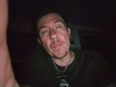 The image shows a close-up of a Caucasian male, seated, wearing a dark green t-shirt and a silver chain necklace with a pendant. The man is looking directly at the camera, with a slight smile on his face. The background is dark and blurred, possibly taken indoors, with a hint of a wall behind him. The coordinates indicate that the image was likely taken in Anderson, South Carolina, USA, specifically near the intersection of North Main Street and West Whitner Street, near the Anderson YMCA.