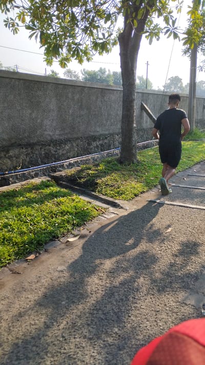 In Sidodadi, Indonesia, a person is captured in a rear-view, mid-stride while jogging on a paved path. The individual, wearing a black t-shirt and shorts, is positioned in the right third of the frame, moving away from the camera and towards the right. Their shadow is cast prominently on the path, extending towards the left.
The setting appears to be an outdoor area with a concrete wall forming a backdrop on the left. A tree stands between the wall and the jogging path, with its branches extending into the upper portion of the image. Adjacent to the wall and path, there is a strip of green grass. A blue pipe runs along the base of the wall, near the grass. The path itself is textured asphalt, with some subtle markings or repairs visible. The lighting suggests it is daytime, possibly morning or late afternoon, given the length and angle of the shadows. The overall environment seems to be a well-maintained urban or suburban outdoor space.