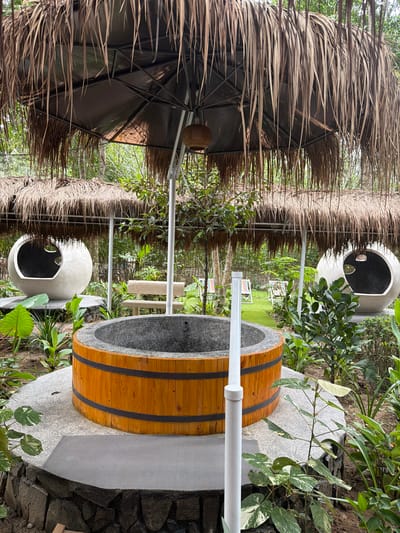 The image depicts an outdoor seating area with a tropical aesthetic, likely located in Suối Câu, Vietnam. In the foreground, a circular, wooden-clad tub with a stone interior sits on a raised, stone-and-concrete platform. A white vertical pipe is situated to the right of the tub. Behind the tub, a thatched roof structure supported by white poles covers the area. To the left and right of the tub are large, spherical, concrete seating pods with cut-out entrances. In the mid-ground, a stone bench is visible, and beyond that, a patch of artificial turf with several colorful lounge chairs. The background is filled with lush green foliage and trees, indicative of a natural or landscaped environment. The scene is serene and suggests a relaxing or recreational space.