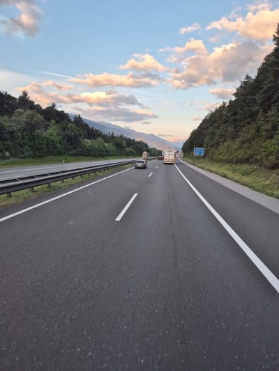 The image captures a view from a vehicle on a highway in Terfens, Austria, at dusk. The highway is a multi-lane asphalt road with clear white lane markings. A metal guardrail separates the highway from the adjacent grassy shoulder and a dense forest. In the distance, mountains are visible, partially obscured by trees and the evening sky. The sky is a gradient of blue and pale orange, with scattered clouds illuminated by the setting sun. Several vehicles are present on the highway, including cars and trucks. One truck in the foreground has a white trailer with "HÜBNER" printed on it. A blue highway sign is visible on the right side of the road, indicating distances to various locations, including Innsbruck. The overall scene depicts a typical highway travel experience in a mountainous, forested region during the evening.