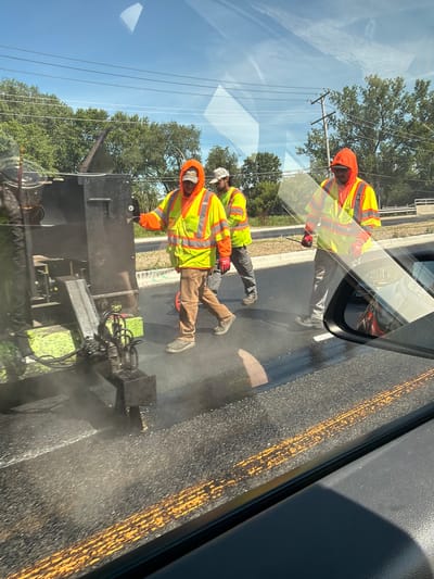 Three male road workers, wearing high-visibility vests over orange hooded sweatshirts and work pants, are performing road maintenance on a sunny day in Buffalo Grove, Illinois, United States of America. One worker on the left is operating a piece of road paving equipment that is emitting steam. Another worker in the center is walking beside the equipment, and a third worker on the right is also walking alongside. The road surface is dark asphalt with fresh orange markings. Trees and power lines are visible in the background, and the scene is framed by reflections on a car's windshield and side mirror.