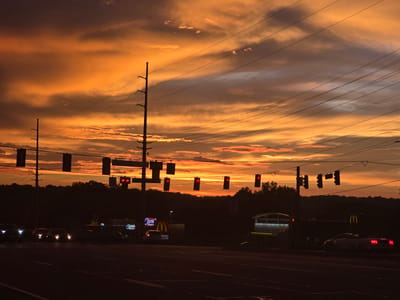 This is a horizontal, medium-angle, outdoor photograph taken at dusk in Woodstock, Georgia, United States of America. The sky is a dramatic display of orange, yellow, and gray clouds, illuminated by the setting sun. Several power lines stretch across the frame from left to right, supported by utility poles. In the middle ground, a tree line forms a dark silhouette against the vibrant sky. Below the trees, the lights of businesses are visible, including a McDonald's with its iconic golden arches and a sign for "Canes." Traffic lights are suspended over the intersection, with several showing red lights. Vehicles are present on the road, with headlights and taillights illuminated. The foreground depicts a dark asphalt road. The overall mood is serene yet dynamic, capturing the transition from day to night in an urban or suburban setting.