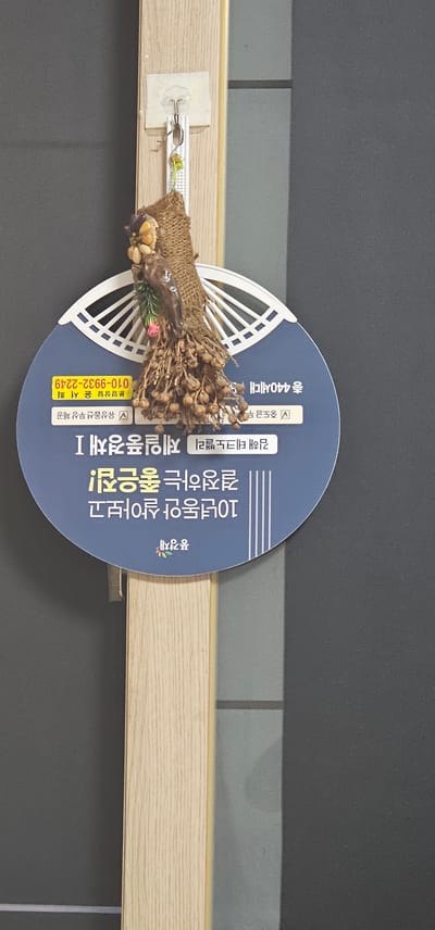 A circular blue sign is attached to a light wood-grained pole against a dark gray background. The sign has white text and a decorative graphic. At the top of the sign, there is a graphic of a fan shape with radiating lines. Below this, a bunch of dried plant material hangs from a burlap wrap, obscuring the sign.
The text on the sign appears to be in Korean. In the upper right, there is text that reads "종 440세대" which translates to "440 households". Below this, in the center, is text that reads "김해시" which translates to "Gimhae City". In the lower part of the sign, there is a logo with some small colorful flowers and the text "김해시" again.
There is a yellow rectangular sticker on the left side of the sign with Korean text and a phone number: "010-9932-2249". This is likely a contact number for Gimhae City.
The sign is hanging from a small clear hook attached to the pole. The overall scene suggests an outdoor public space in Gimhae City, South Korea.