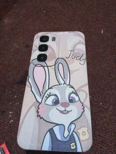 A phone case rests on a brown textured surface. The case features a cartoon illustration of Judy Hopps from Zootopia. Judy's character is depicted in her police uniform, with her signature bunny ears and bright eyes. The name "Judy" is written in cursive above her head. The camera cutouts are visible at the top of the case. The overall color scheme is muted and pastel, with pink and gray tones.