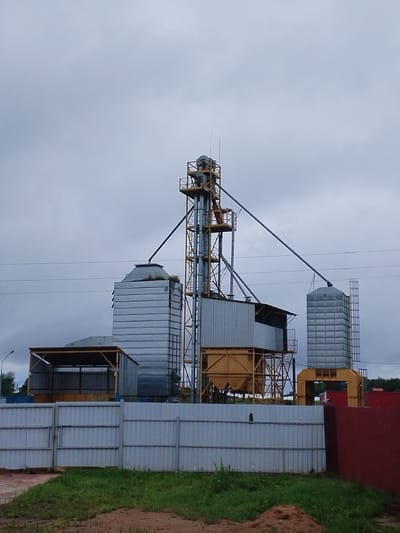 This is a full-shot, eye-level, outdoor, medium-angle photograph taken on an overcast day in the village of Varantsy, Grodno Region, Belarus. The image features a grain elevator complex with several tall, metallic silos and a central processing tower. The silos are cylindrical and ribbed, with one appearing to have a small amount of vegetation growing on its roof. The processing tower has a yellow metal frame structure with various pipes and chutes. In the foreground, a light grey metal fence runs horizontally across the image, separating the viewer from the industrial complex. Beyond the fence, a strip of green grass and some dirt are visible. To the right of the fence, a portion of a dark red metal fence is present. The sky is uniformly grey and cloudy. There are no people or animals visible in the frame. Electrical wires are strung across the upper portion of the image.