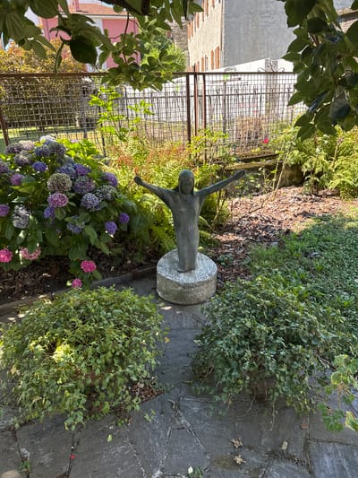 The image depicts a serene garden scene, likely in the city of Domodossola, Italy, given the architectural style of the buildings visible in the background. At the center of the frame stands a bronze statue of a figure with outstretched arms, emerging from a circular stone base. This statue is surrounded by lush greenery, including vibrant hydrangeas in shades of blue, purple, and pink, as well as ferns and other leafy plants. Two large, rounded bushes of ivy flank the statue on the left and right, adding to the verdant atmosphere. The ground is paved with irregular stone slabs, with patches of grass and fallen leaves scattered about. In the background, a metal fence separates the garden from a building with a pink facade and terracotta roof, and a larger, unpainted building with shuttered windows. Sunlight filters through the leaves of overhanging trees, casting dappled shadows on the scene. The overall impression is one of peaceful contemplation and natural beauty.