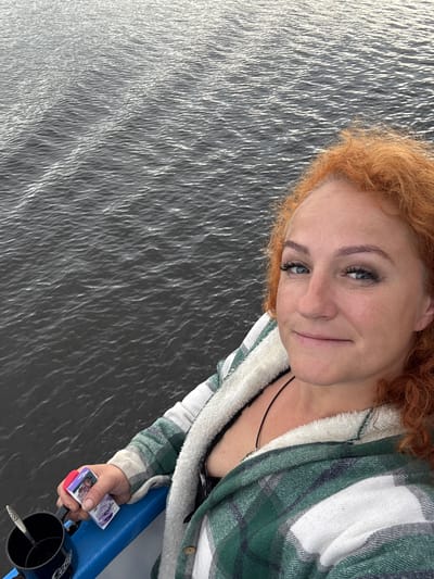 The image shows a woman with red hair, taking a selfie on a boat. She is wearing a green and white plaid jacket with a fuzzy white lining. She is holding a small package in her left hand, which appears to be a pack of cigarettes. A dark blue mug with a spoon in it is visible next to her hand. The background is composed of dark, rippling water. The location is not specified in the image, and there is no information to suggest it is in Zuevo, Russia.