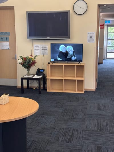 This image captures an indoor common area or waiting room, likely within a facility such as a medical center or care home in Carrum Downs, Australia. The room features beige walls and a dark grey patterned carpet.

On the wall, two flat-screen televisions are mounted. The larger TV on the left is off or displaying static, while the smaller TV on the right shows a still image of two older individuals with white hair, possibly men, embracing or hugging. Above this TV, a "MediStays" logo and a QR code are visible. Below it, several pieces of paper are affixed, some appearing to be informational notices.

To the left of the televisions, a circular wall clock indicates approximately 9:05. A light wooden bookshelf with six empty compartments stands next to the smaller TV. Below this, a small black side table holds a black landline phone and a clear vase containing a colorful bouquet of red, white, and green flowers and foliage. Part of a light wooden, oval-shaped table with a woven coaster is visible in the bottom right corner, suggesting a seating area.

Several signs are present: "FIRE HOSE REEL" in red text near a door frame, and on a wooden door to the bottom left, "SMOKE DOOR DO NOT OBSTRUCT DO NOT KEEP OPEN" and "Staff Toilet" accompanied by gender symbols. A large window or archway in the background reveals outdoor greenery and natural light, indicating it is daytime. The overall impression is a clean, functional, and well-maintained interior.
