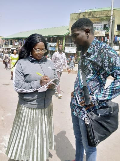 The image depicts an outdoor scene in Mumbwa, Zambia, featuring two individuals in the foreground engaged in an interaction on an unpaved surface, with a bustling commercial street and buildings in the background under a clear sky. On the left, a female subject with long dark hair, wearing a grey zip-up jacket with white stripes on the sleeves (featuring an Adidas logo on the left chest) and a long, light olive-green pleated skirt, holds a small white notebook in her left hand and a yellow pen in her right, actively writing or taking notes. She wears light-rimmed round glasses and looks down at the notebook. On the right, a male subject with short dark hair and a beard, dressed in a long-sleeved shirt with a blue, black, and white geometric pattern and blue jeans, stands facing the female, with his right hand on his hip. He carries a black shoulder bag slung across his body. In the mid-ground, a multi-story building with a green corrugated metal roof and a weathered, light-colored facade houses multiple ground-level shops; visible signage includes "DESIGNERS WARDROBES" and "ENNYFRANK". Several other pedestrians are present in the background, walking along the street.