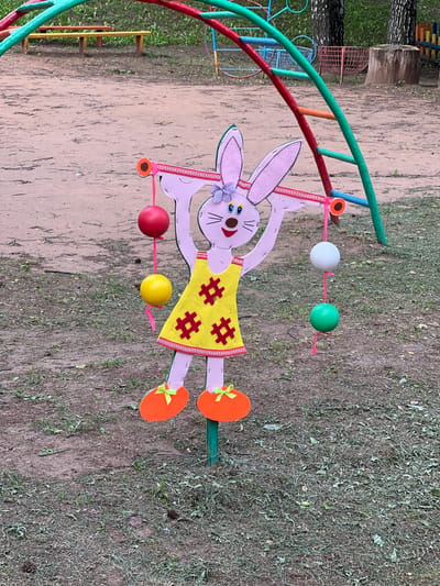 In the city of Советско-Никольское, Russia, a pink cutout of a cartoon bunny stands as a decorative element on a playground. The bunny, adorned in a yellow dress with red hash symbols, is positioned outdoors on a dirt and grass ground. It holds a pink ribbon in its outstretched arms, from which red, yellow, white, and green balls are suspended. The bunny's feet are represented by orange ovals with green bows. In the background, a colorful jungle gym structure with green, red, and blue bars dominates the scene. Beyond the playground equipment, a few trees with white trunks are visible, along with a wooden stump and a short fence. The ground is a mix of sandy soil and sparse, dry grass, with some scattered pinecones.