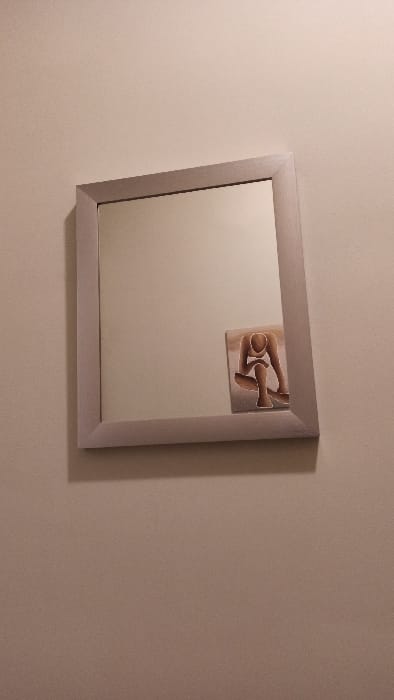 The image displays a rectangular mirror mounted on a plain, light beige wall in Florenville, Belgium. The mirror features a light gray, angular frame. Reflected within the mirror, positioned in the lower right quadrant, is a small rectangular artwork. This artwork depicts a stylized, faceless human figure with light brown skin tone and white outlines, rendered in a crouched or seated posture with knees bent and the body leaning forward. The wall itself shows no other distinguishing features or objects.