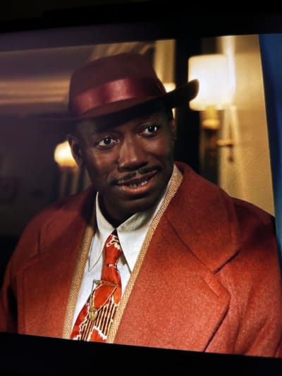 A man wearing a burgundy fedora and a rust-colored coat is shown from the chest up, looking to his right. He has dark skin, a mustache, and is smiling. He is wearing a white shirt, a patterned tie with orange, white, and brown elements, and a tie clip. The coat has a contrasting trim along the lapels. In the background, a lamp with a white shade and a gold base is visible, along with a hint of what appears to be a wooden railing or trim. The image is a medium close-up shot, with the lighting creating a warm glow. There are no discernible elements that would place this image in Dongaron, Russia.