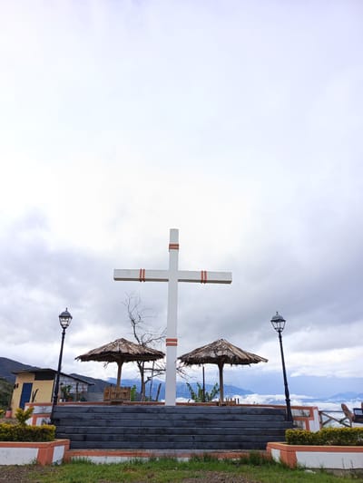 The image shows a large white Christian cross with red stripes at the top and center, situated on a platform in the city of El Cisne, Ecuador. The platform is made of gray concrete steps bordered by white walls with orange trim. Two thatched-roof structures with benches sit on the platform behind the cross. Two black street lamps stand on either side of the platform. Behind the platform, a small yellow building with a blue door and a shack are visible. A mountain range is visible in the distance, obscured by clouds. The sky is overcast. The ground in front of the platform is covered in green grass.