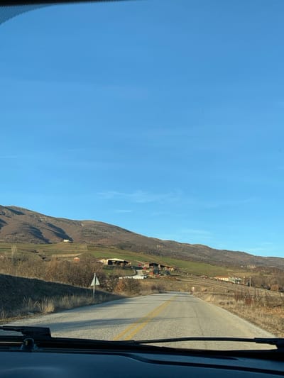 The image captures a wide, sunny landscape from the perspective of a vehicle, likely looking out a passenger-side window. The scene is dominated by a vast, clear blue sky with a few faint, wispy clouds, occupying the top half of the frame.

In the lower half, a rugged mountain range stretches across the left, its slopes covered in brownish, sparse vegetation, suggesting a dry season or early spring. Below the higher slopes, patches of green fields and scattered clusters of small, rural buildings, possibly homes or farm structures, are visible in a valley or lower plain.

A paved road runs along the right side of the frame, curving gently into the distance. It features a prominent double yellow line down its center, indicating traffic regulations. The dark interior of the vehicle, including what appears to be a door frame and a windshield wiper, is visible on the far right and bottom right edges, framing the view. A white, arrow-shaped road sign, pointing left, stands on the roadside.

The bright daylight suggests it is either morning or afternoon, with clear weather. The overall impression is one of a tranquil, scenic drive through a mountainous, rural area. Given the context of Grevena, Greece, the landscape aligns with the region's known mountainous terrain and rural settlements. No people are visible in the image.