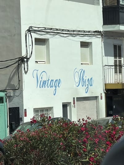 The image shows the exterior of a white building with blue lettering that reads "Vintage Ibiza". The building has two windows on the upper floor and a garage door on the lower floor. There are electrical wires running along the side of the building. In front of the building, there is a bush with red flowers and a car. The building appears to be in a commercial or residential area. The image is taken from a slightly low angle, looking up at the building.