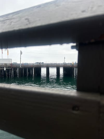 The image displays Stearns Wharf in Santa Barbara, United States, viewed through a rectangular opening in a dark, weathered wooden structure. The foreground consists of dark wooden planks framing the scene. A wooden pier extends horizontally across the midground, supported by numerous dark vertical piles submerged in dark green, rippled water. On the pier, vehicles, including a white box truck and at least two cars, are visible. Utility poles with strung wires run along the pier's length. On the left, an American flag and a yellow flag are present near a white structure. On the right side of the pier, a small group of approximately three to four people is observed, with one individual wearing a yellow top and another a blue top. The sky is overcast and uniformly light gray. In the distant background, a marina with numerous sailboat masts is discernible.