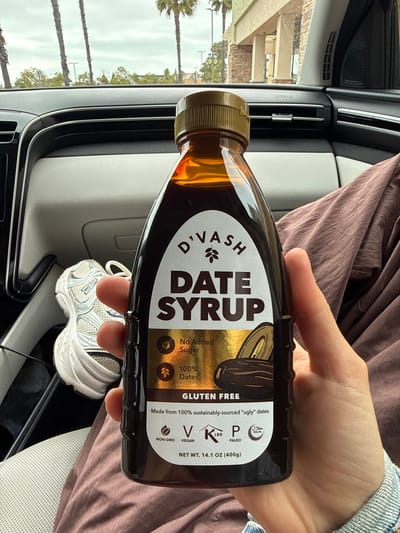 A light-skinned human hand holds a 14.1 oz (400g) bottle of D'VASH Date Syrup, filled with a dark brown liquid and sealed with a golden-brown ribbed cap. The bottle's label is predominantly white and gold, prominently displaying "D'VASH DATE SYRUP" and listing attributes such as "No Added Sugar," "100% Dates," "Gluten Free," and "Made from 100% sustainably-sourced "ugly" dates." Certifications for NON-GMO, VEGAN, KETO, PALEO, and HALAL are present at the bottom of the label. The hand is positioned over the individual's lap, which is covered by a reddish-brown, textured fabric. The right wrist exhibits a light blue denim cuff.

The immediate environment is the interior of a vehicle, viewed from the perspective of the passenger seat. The car's dashboard features black and silver/grey trim, with an "AIRBAG" inscription visible on the passenger side and air vents present. A white athletic shoe, characterized by mesh and synthetic overlays, rests on the floor or passenger seat to the left of the bottle.

Through the front windshield, an outdoor scene unfolds under an overcast sky. Several tall palm trees are visible, indicative of a warm climate. Below the tree line, a multi-story commercial building is present, featuring prominent light-colored pillars and a textured, possibly stone or stucco, facade on its lower sections. This architectural style, combined with the presence of palm trees, points to a commercial parking lot setting, characteristic of a shopping center or strip mall within the city of Oceanside, California.