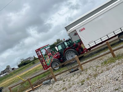 The scene is set in Curtistown, Ireland, under an overcast sky. A red agricultural tractor, fitted with a green-tinted cabin and an orange rooftop beacon, is positioned on a gravel surface. Attached to the rear of the tractor is a large, red agricultural implement featuring multiple vertical arrays of green, cup-shaped components. To the right of the tractor, a white trailer or lorry body is present, displaying "VHS" in red lettering with an associated logo, and "WSD Farm Machinery" in smaller print. The trailer's side curtain is secured by black straps. A three-rail wooden fence spans the mid-ground, separating the gravel area from a grassy field. Miscellaneous items, including a roll of wire fencing and a partially visible blue object, are on the gravel near the fence. A yellow rope or cable lies on the ground. In the background, beyond the grassy field, several buildings with dark roofs are visible amidst trees.