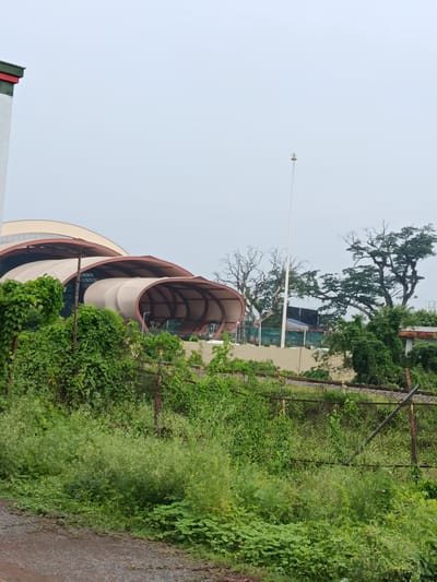 The image captures a scene in Benapole, Bangladesh, featuring a large, modern building with a distinctive curved, canopy-like roof structure. The building appears to be under construction or newly built, with some areas showing exposed structural elements and sheeting. Lush green vegetation, including trees and dense bushes, surrounds the building, with a rusty wire fence running horizontally in the foreground. A dirt road is visible on the lower left side of the image. The sky is overcast and a tall, thin pole stands in front of the building, extending towards the sky.