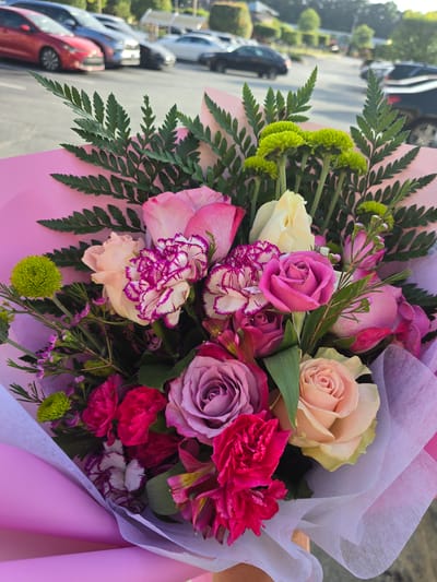 A close-up, vertical shot shows a bouquet of flowers wrapped in pink and white paper. The bouquet contains a variety of flowers including roses in shades of pink, purple, and peach, as well as white and pink carnations with purple edges, and small green chrysanthemums. The background is blurred and shows a parking lot with several cars, including a red sedan and a black SUV, suggesting an outdoor location, possibly a commercial area or residential street. The lighting suggests it is daytime, with some sunlight visible. Based on the context provided, the location is the city of Duluth, United States of America.
