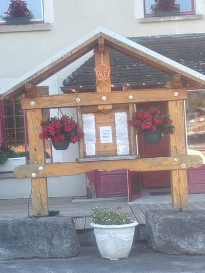 The image shows a wooden stand with two hanging flower baskets filled with vibrant red flowers. The stand is located outdoors, in front of a building with a red door. A sign that reads "OUVERT" is attached to the stand, suggesting the building is open for business. The scene appears to be in a small town or village, possibly in a rural area. The overall impression is one of a charming and welcoming establishment. The location in Charchilla, France, adds a layer of specificity to the scene, suggesting a picturesque European setting.