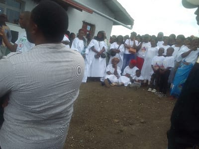 A crowd of people gathers outdoors, many dressed in white garments. Some individuals are seated on the ground, including children. A man with a striped shirt stands in the foreground, partially obscuring the view. Other individuals wear t-shirts with a design on them. The background features a light-colored building with windows and a gutter. The ground appears to be dirt or bare soil.