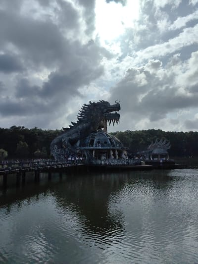 The image depicts the abandoned Ho Thuy Tien water park in Kim Long Ward, Hue, Vietnam. A large, weathered concrete dragon sculpture forms the central subject, its body intricately wrapped around a multi-level, dilapidated domed building situated within a dark body of water. The dragon exhibits prominent scales, spiky features along its back, and an open mouth revealing sharp teeth and horns. The underlying domed structure features arched openings and pillars, showing signs of decay and environmental wear.
A concrete walkway with railings extends from the left side of the frame, supported by pillars in the water, leading towards the base of the main dragon structure. To the right of the primary dragon, a smaller, weathered domed structure topped with a decorative element resembling a dragon's head or fin is also present in the water.
Several individuals are visible on the walkway and near the entrance of the main dragon building, appearing as visitors or explorers. The background consists of a dense line of dark green trees and foliage. The sky is predominantly overcast with heavy grey clouds, though a bright area with visible sun glare is present in the upper right quadrant. The surface of the body of water exhibits minor ripples and provides reflections of the structures and sky.