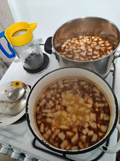 The image shows a stove top with two pots of stew. One pot is stainless steel, and the other is white with a black rim. Both contain a stew with a brownish broth and cubed white vegetables, possibly potatoes or turnips. To the left is a glass pitcher with a yellow lid and blue handle. There is a plate with silverware next to the pot. The image could be from a kitchen in Musachevo, Bulgaria, but there is no confirmation.