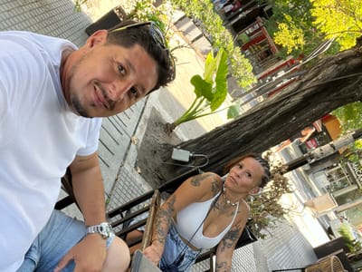Two individuals are depicted in an outdoor setting in City Bell, Argentina. A male subject occupies the left foreground, presenting light skin, short dark hair, and stubble. He wears a white t-shirt, light shorts, and a silver wristwatch on his left wrist, with sunglasses resting on his head. He smiles directly at the camera. To his right, a female subject, also with light skin, has braided hair and multiple tattoos visible on her arms and chest. She wears a white halter top, denim shorts, and a shell necklace, looking towards the camera with a neutral expression. Both are seated in dark-colored metal chairs on a paved sidewalk. A large tree with green foliage stands between the subjects and the background, where a street, partial vehicles, and commercial buildings with visible signage, including "MACAR...", are present under natural daylight.