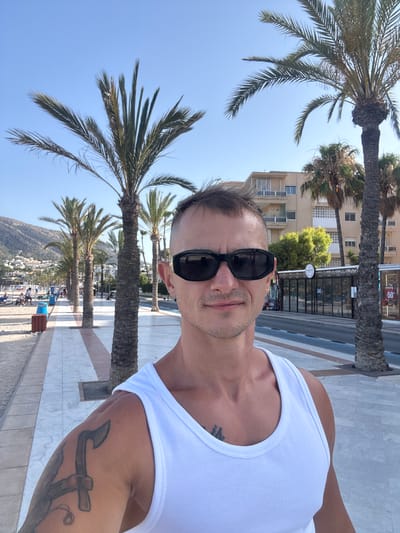 A man wearing a white tank top and sunglasses takes a selfie on a sunny day in l'Alfàs del Pi, Spain. He is standing on a paved walkway lined with palm trees, with a sandy beach to his left and buildings and a road to his right. The man has a tattoo on his left arm. In the background, there are buildings, more palm trees, and a hill with houses. The sky is clear and blue.