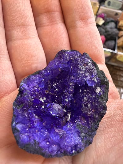 An adult human hand, exhibiting light skin tone and visible palm and finger creases, holds a roughly palm-sized, irregular purple mineral geode in an open-palm presentation. The geode features a central cavity densely lined with small, reflective, deep purple crystalline structures. A few larger, faceted crystal points are observable within the cluster. The outer perimeter of the geode consists of a dark, rough rock matrix. The background is out of focus, showing indistinct forms and colors consistent with a collection or display of other mineral or stone objects. The scene is located in the city of Arucas, Spain.