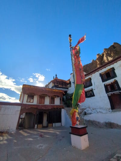 The image depicts a Buddhist monastery in Yulang, India. The foreground features a prayer flag pole with colorful prayer flags fluttering in the wind. In the background, traditional white-washed monastery buildings with wooden window frames and roofs are visible, nestled against a rocky hillside under a clear blue sky with scattered clouds. The architecture suggests a remote, mountainous region. The lighting indicates it is daytime, with sunlight casting shadows on the ground.