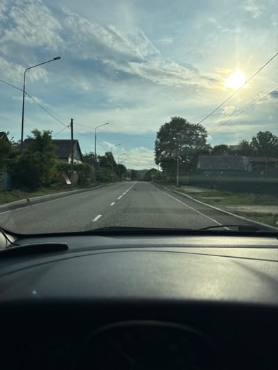 The image depicts a view from inside a vehicle looking out at a rural road in Kurinskaya, Russia. The sun is bright and high in the sky, casting rays through wispy clouds, suggesting it is daytime, likely midday or early afternoon. The weather appears clear and sunny.

The road itself is paved and has a white dashed line marking the center. On the left side of the road, there are houses with pitched roofs, surrounded by trees and greenery. Streetlights line the side of the road. Power lines crisscross the sky. The right side of the image is dominated by the interior of the vehicle, showing the dashboard and windshield. The perspective suggests the vehicle is in motion, traveling along the road.

There is no visible text in the image. The overall mood is peaceful and serene, characteristic of a sunny day in a rural setting. The scene captures a moment of travel through a quiet, residential area.