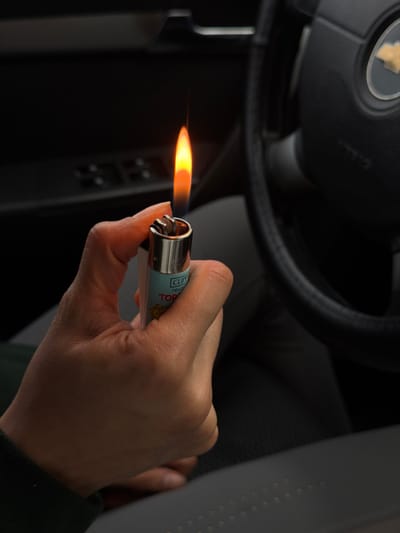 A person holds a lit Clipper lighter inside a car. The flame burns horizontally from the lighter's metal nozzle. The person's hand and wrist are visible, with a dark sleeve suggesting the person is wearing a jacket or long-sleeved shirt. In the background, part of the car's steering wheel, dashboard, and seat can be seen, indicating the setting is the car's interior. The overall scene has a dark ambiance.