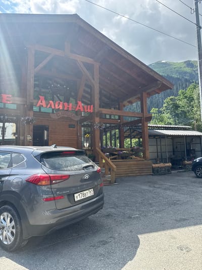 This media file depicts an outdoor scene at a cafe in Dombay, Russia. The central subject is a wooden building with a prominent A-frame roof and visible timber framing. Large red lettering spells out "Алан-Аш" on the facade, indicating the name of the establishment, likely a cafe or restaurant. A sign above an entrance reads "ОТКРЫТО" (Open).

In the foreground, the rear of a dark grey Hyundai Tucson is parked on an asphalt surface. The car's license plate, "Y 178BT 761 RUS," is clearly visible. The setting appears to be mountainous, with lush green trees and a cloudy sky. The sun is shining, casting a shadow from the car onto the pavement. The overall impression is one of a bright, possibly summer day in a scenic, mountainous region. There are no people visible in the image, and the activity seems to be centered around the cafe as a destination. The building's rustic wooden architecture complements the natural mountain environment.