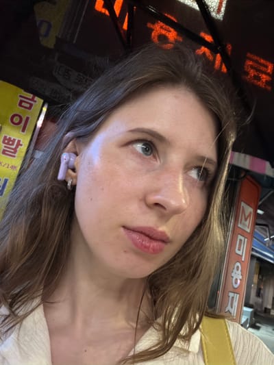 A close-up, indoor portrait captures a young woman with light skin and blue eyes, looking to her right. She has medium-brown hair that falls around her face and neck. A light purple earbud is visible in her right ear, accompanied by a small hoop earring. She is wearing a cream-colored collared shirt. In the background, to the left, a bright yellow sign with Korean text is partially visible. To the right, an orange sign with Korean text, reading "미용재" (miyongjae, meaning "cosmetic material" or "hairdressing material"), is also visible. The overall lighting suggests an urban evening setting, possibly a street in Uijeongbu-si, South Korea, given the presence of Korean signage.
