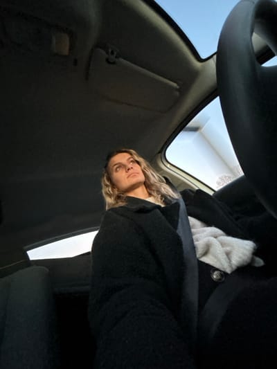 A woman with blonde wavy hair is captured from a low angle inside a car. The steering wheel dominates the upper part of the frame. The woman is wearing a dark, textured coat and a light-colored scarf. The car's interior features a sun visor, windshield, and side window. Light enters through the windshield, creating a contrast within the car's cabin.