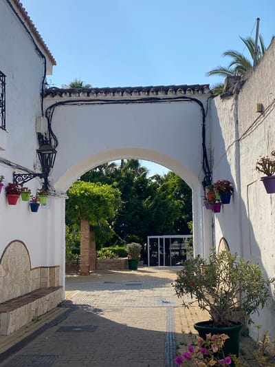 This is an outdoor shot in Cancelada, Spain, featuring a white arched passageway leading to a courtyard. The passageway is framed by white stucco walls, one of which displays several hanging potted plants in various colors, including pink, red, green, and blue. A vintage black lantern is mounted on the wall to the left of the archway. The courtyard is paved with interlocking bricks and features a stone bench on the left. Lush green trees and palm fronds are visible through the archway, and a large potted plant with pink flowers sits on the right side of the paved area. The sky is clear and blue.