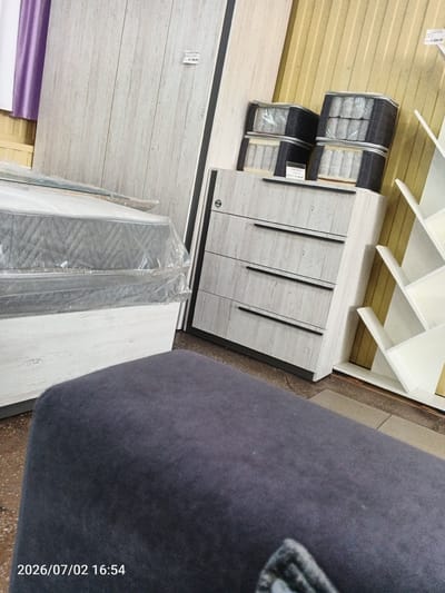 The image depicts an indoor scene, likely a furniture store, with several pieces of furniture on display. In the foreground, a dark gray upholstered piece of furniture, possibly a sofa or bench, dominates the bottom half of the frame. The top left of the image shows a portion of a room with a lavender curtain.
The middle ground features a large, white wooden bed frame with a mattress covered in plastic. Next to the bed, to the right, is a light-colored wooden dresser with four drawers, each adorned with a black handle. On top of the dresser are stacks of packaged mattresses, also wrapped in plastic. To the right of the dresser, a white bookshelf with an angular design is visible. The background consists of a yellow, corrugated metal wall.
A price tag is partially visible on the dresser, and another price tag is attached to the wall behind the bed. The bottom left corner of the image has a timestamp: "2026/07/02 16:54". While the image provides visual information about the scene, it does not contain any explicit details that would allow for the determination of its location in the city of Chany, Russia.