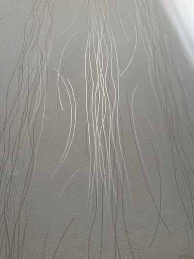 The image shows a close-up of textured wallpaper with a pattern of thin, vertical, wavy lines. The wallpaper is a light gray color. The texture appears to be a raised, stippled effect. The wavy lines are a slightly lighter shade of gray or silver and are subtly raised or have a sheen that catches the light, creating a subtle contrast with the background. The lighting suggests a light source from the top right, casting a slight gradient across the surface and highlighting the texture and lines. There are no people, animals, or specific objects visible other than the wallpaper itself. The location in Anadyr, Russia, is not discernible from the image, as it depicts a generic interior surface.