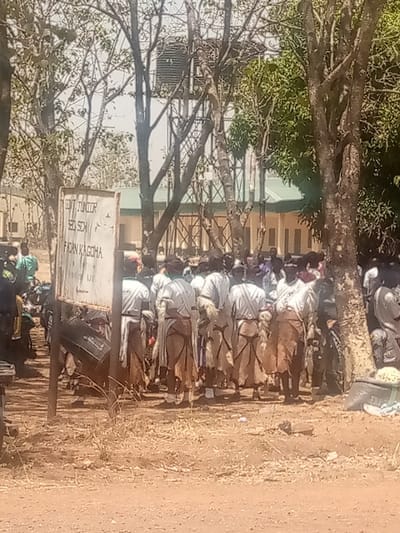 The image depicts an outdoor scene at the Government Junior Secondary School, Fadan Kagoma, Kagoma, Nigeria. A prominent white sign in the left foreground displays the text "GOVT JUNIOR SED SCH FADAN KAGOMA". A group of individuals, primarily observed from behind, stands in the central and right midground. They are dressed in light-colored tops and brown, wrap-around lower garments, with some exhibiting fur-like attachments on their backs. The ground surface is dry, reddish-brown earth. To the left of the sign, several motorcycles are visible. In the background, partially obscured by numerous trees with sparse foliage, are buildings with a green roof and a metal water tower supporting a black tank. The scene is illuminated by bright ambient light.