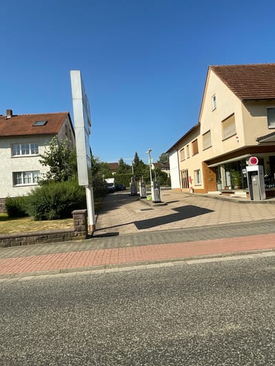 The image captures a view of a street in Wartenberg, Germany, on a clear, sunny day. In the foreground, a two-lane asphalt road is visible, with a sidewalk made of red brick pavers and gray concrete curbing separating it from the properties.

To the left, a two-story residential building with a red tiled roof and white-framed windows stands behind a low stone wall and a well-maintained lawn with green bushes. A tall, white signpost is positioned in front of this building.

To the right, a commercial building with a light yellow facade and a brown tiled roof dominates the scene. This building features large glass windows, a covered entrance, and several smaller windows with what appear to be rolling shutters. Attached to the front of this building is a sign with "LUCKY STRIKE" prominently displayed in red. Adjacent to this sign are modern gas pumps, suggesting the building may house a gas station or convenience store.

Several gas pumps are also visible further down the paved area, with one or two vehicles partially visible in the background, indicating ongoing activity. Greenery, including trees and bushes, is scattered throughout the scene, contributing to a suburban or town-like atmosphere. The overall impression is a well-kept, sunny street in a German town.