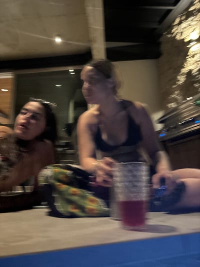 The image is a blurry shot of two young women sitting at a table, taken inside a dimly lit room. One woman, with long dark hair, is sitting to the left of the frame and is out of focus. She is wearing a white tank top. The other woman, with long dark hair, is seated to the right of the frame and is in slightly sharper focus, wearing a black sports bra. Her right hand is holding a glass of red liquid, while her left hand rests on the table. There is a bag with sunflower patterns and other unknown objects on the table. The background is out of focus and shows a brick wall, a glass door with lights, and a stainless steel appliance. The location is in Santa Fe, Costa Rica (latitude: 9.647, longitude: -85.166).