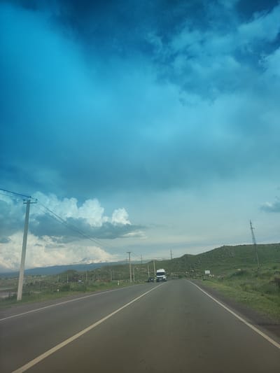 A view from a vehicle driving on a paved road towards a distant mountain range under a dramatic, cloud-filled sky. The road has a white dashed line marking the center. To the left of the road, there are utility poles with wires and grassy fields. To the right, the landscape is characterized by rolling green hills dotted with rocks and sparse vegetation. In the distance, snow-capped mountains are visible. A white truck and a few cars are on the road ahead, and a small herd of animals, possibly sheep, can be seen grazing on the right hillside. The sky is a vibrant blue, transitioning to darker, stormier clouds on the left. The scene appears to be in or near Horom, Armenia, given the characteristic landscape and mountain views.