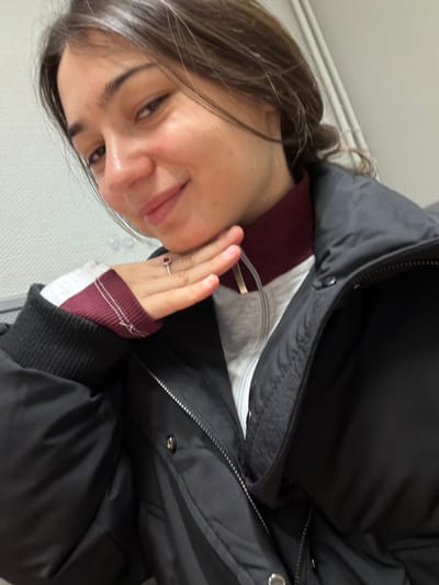 A woman with long brown hair smiles and holds up her left hand, showing off a ring with a red stone. She's wearing a black winter coat over a white top with a maroon collar and cuffs. The coat's zipper is partially open, revealing a silver zipper and snaps. The background appears to be a plain wall, possibly indoors. The image has been rotated counterclockwise.